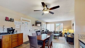 Un comedor con una mesa y sillas de madera, un ventilador de techo y una zona de cocina con armarios y electrodomésticos.