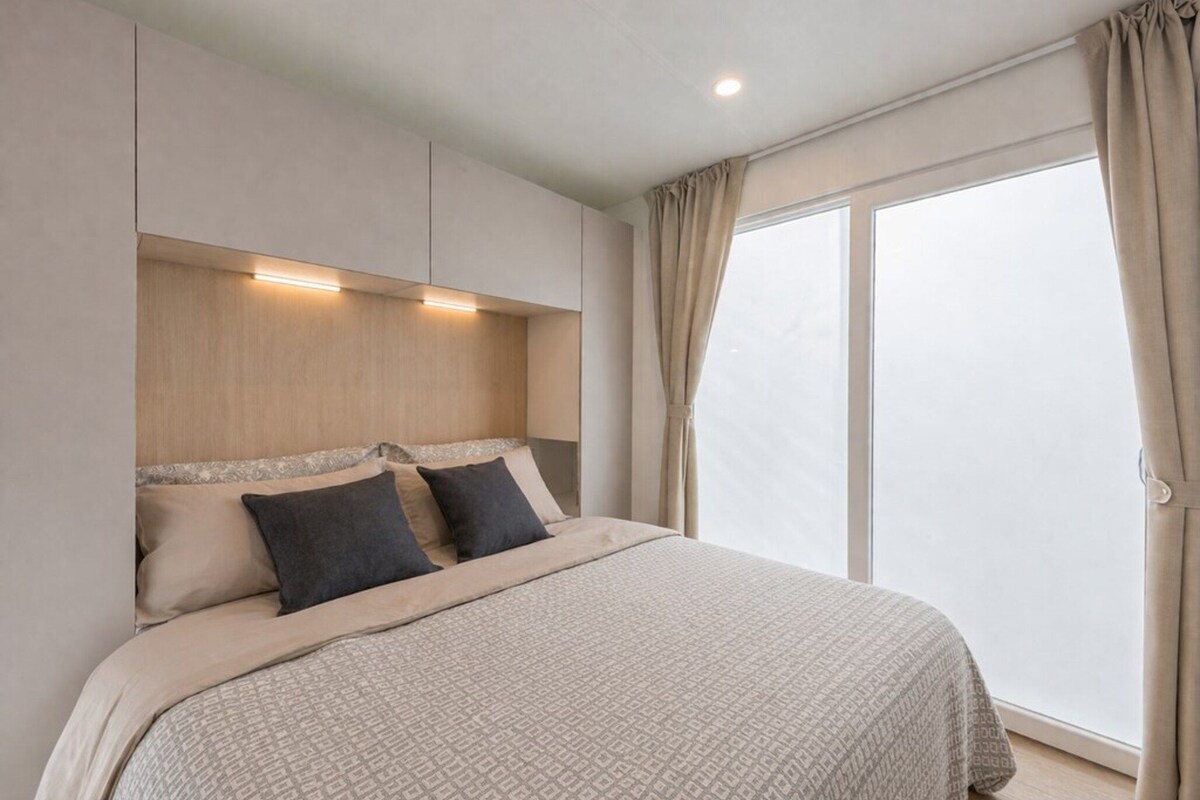 A bedroom with a large bed, a headboard with built-in lighting, and a view through a window with curtains.