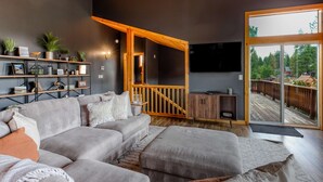 Uma sala de estar moderna com um sofá cinzento, escada de madeira e uma janela ampla com vista para um terraço.