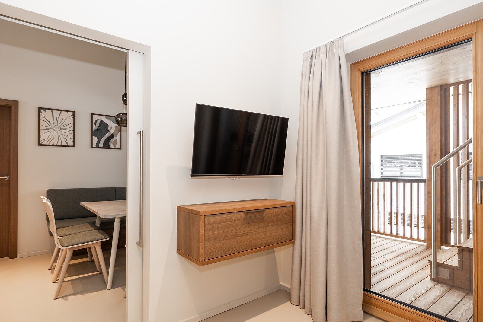 Ein modernes Zimmer mit einem wandmontierten Flachbildfernseher, einem Holzschrank darunter, einem Esstisch mit Stühlen und einer Glastür zum Außenbalkon.