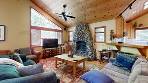 A cozy living room with a stone fireplace, wooden ceiling, and large windows. - Lake Tahoe Cabin with Hot Tub, Private Studio & Yard - Near Resorts (Tahoe City)