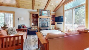 L’intérieur chaleureux d’un chalet avec un plafond en bois, une cheminée en pierre, un canapé, un banc et un téléviseur.