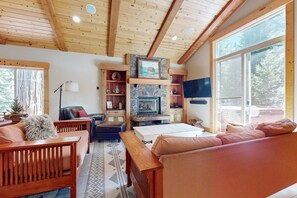 Interior acogedor de una cabaña con techo de madera, chimenea de piedra, sofá, banca y televisor.