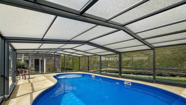 A covered swimming pool with a glass ceiling, a patio area with chairs, and a view of the outdoors.