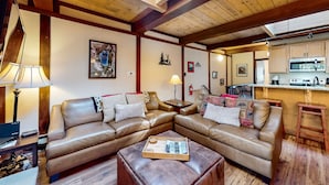 Una sala de estar acogedora con techo de madera, un conjunto de sofá, una mesa de centro y una cocina visible al fondo.