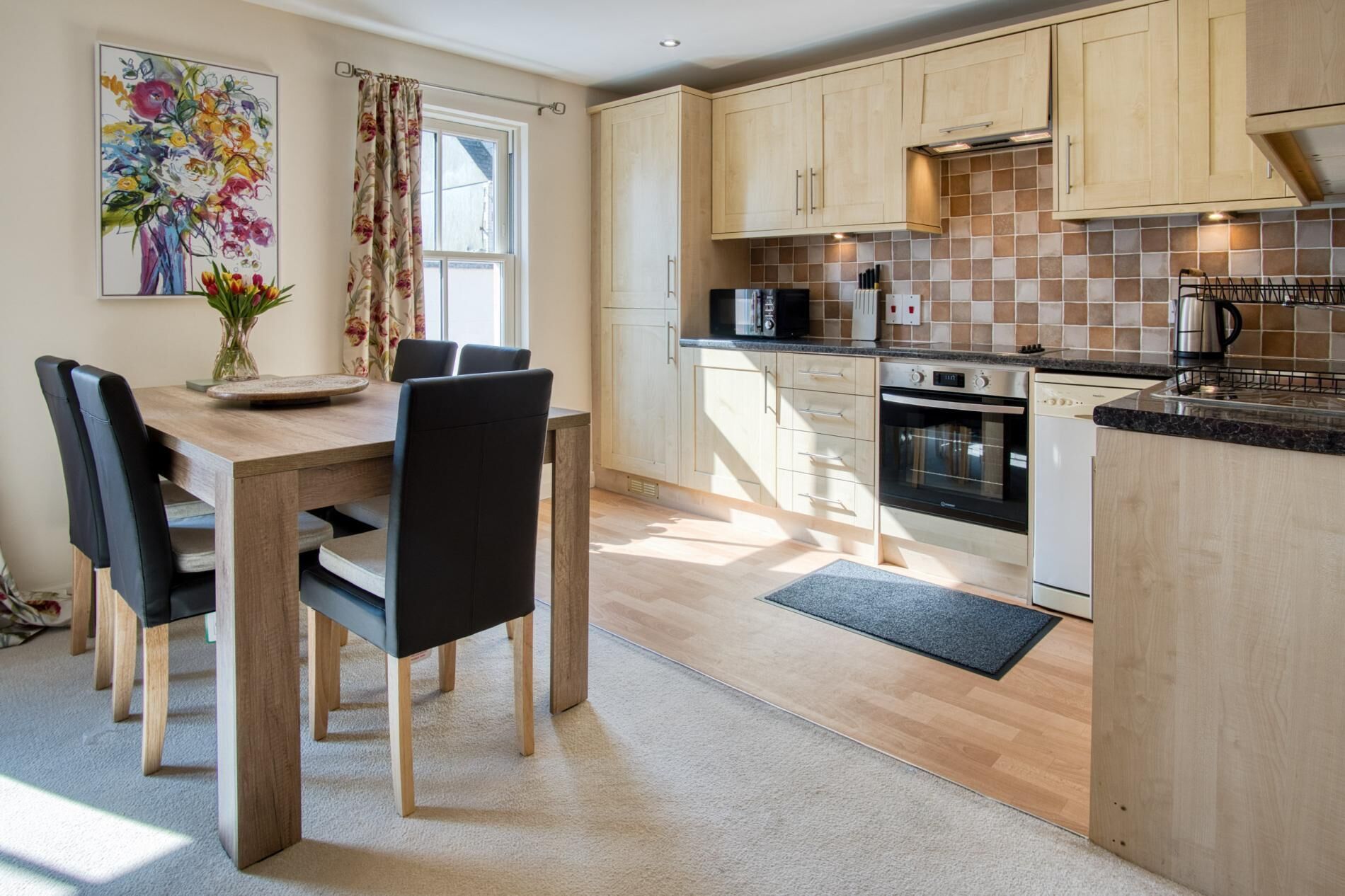 A modern kitchen with wooden cabinets, a dining table with chairs, and a large window with curtains.