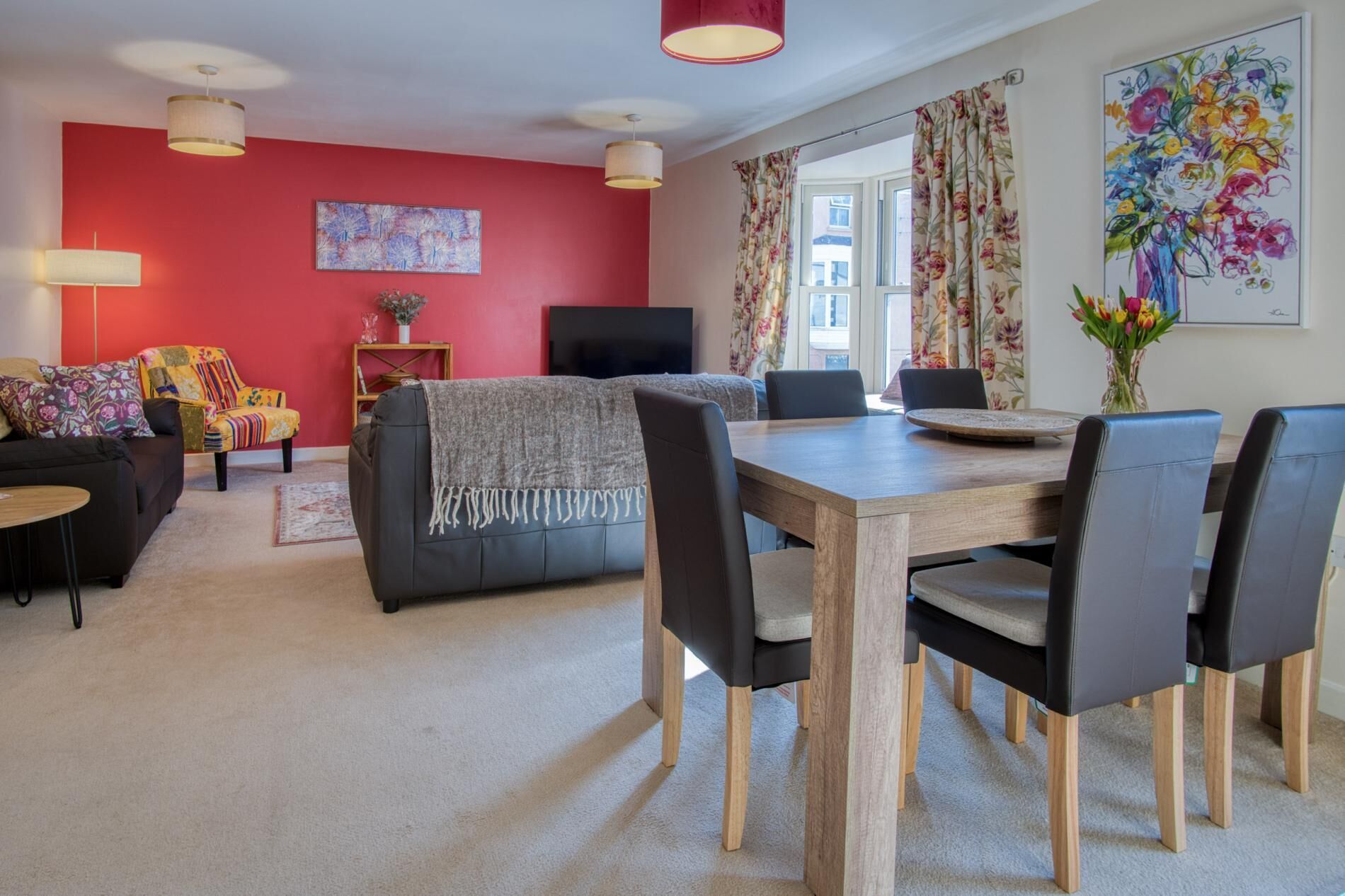 Un salon moderne avec un mur d’accent rouge, un canapé noir, une table à manger en bois avec des chaises et un téléviseur.
