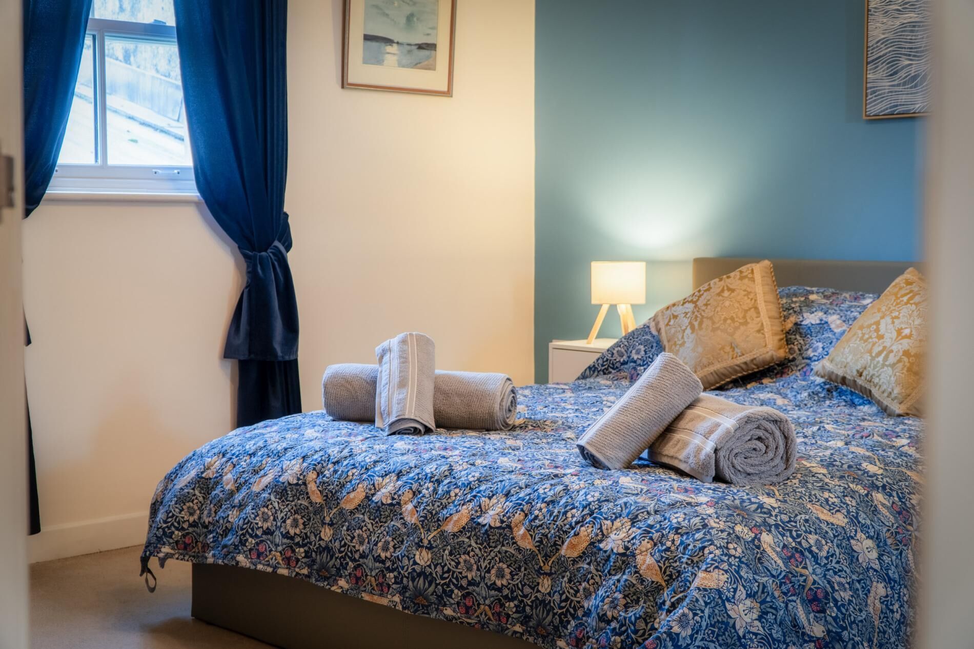 Ein Schlafzimmer mit einem Bett, Kissen, einem Nachttisch, einer Lampe und einem Fenster mit Vorhängen.