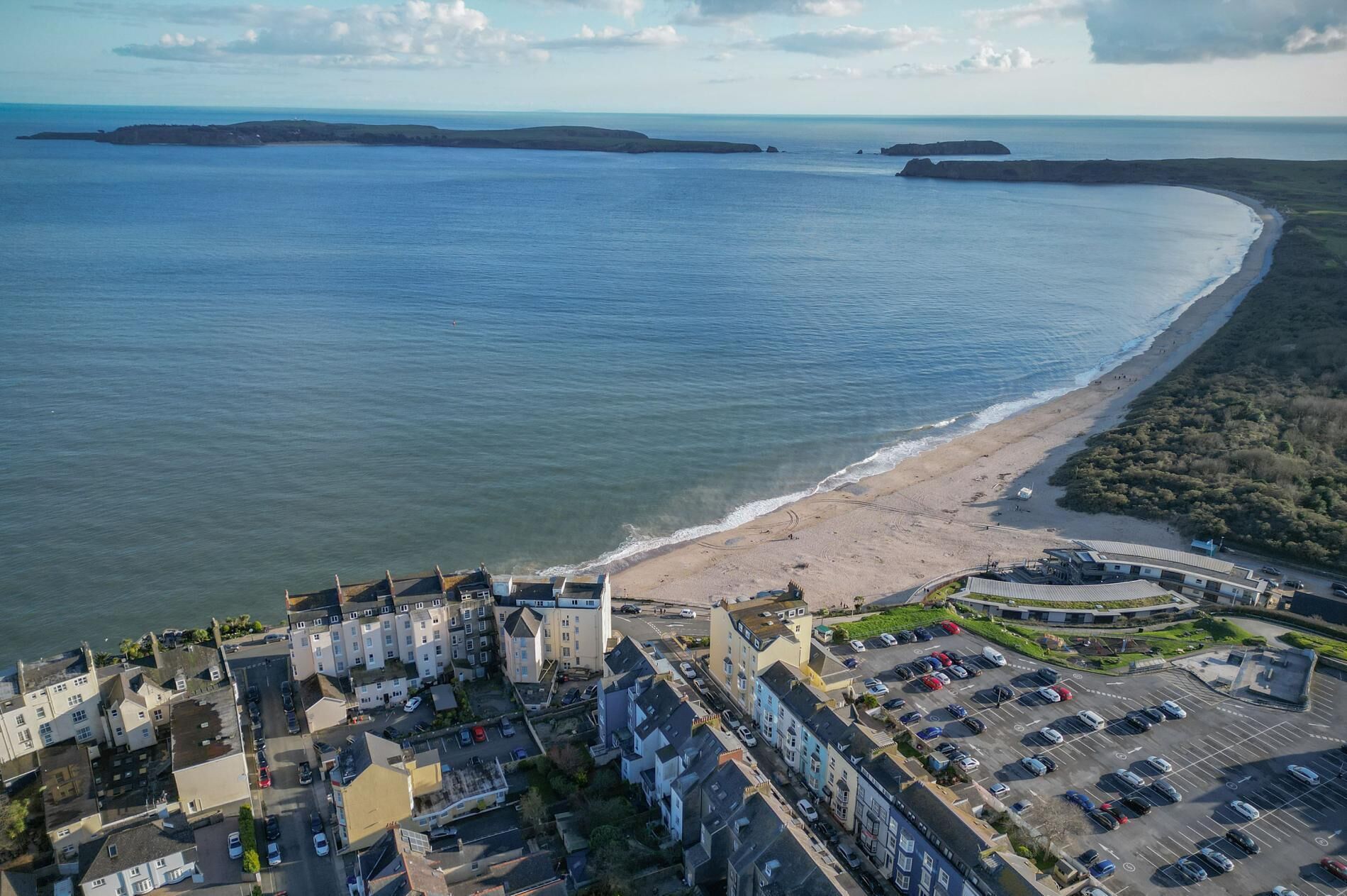 An aerial view of a coastal town with a beach, residential buildings, and a parking area.