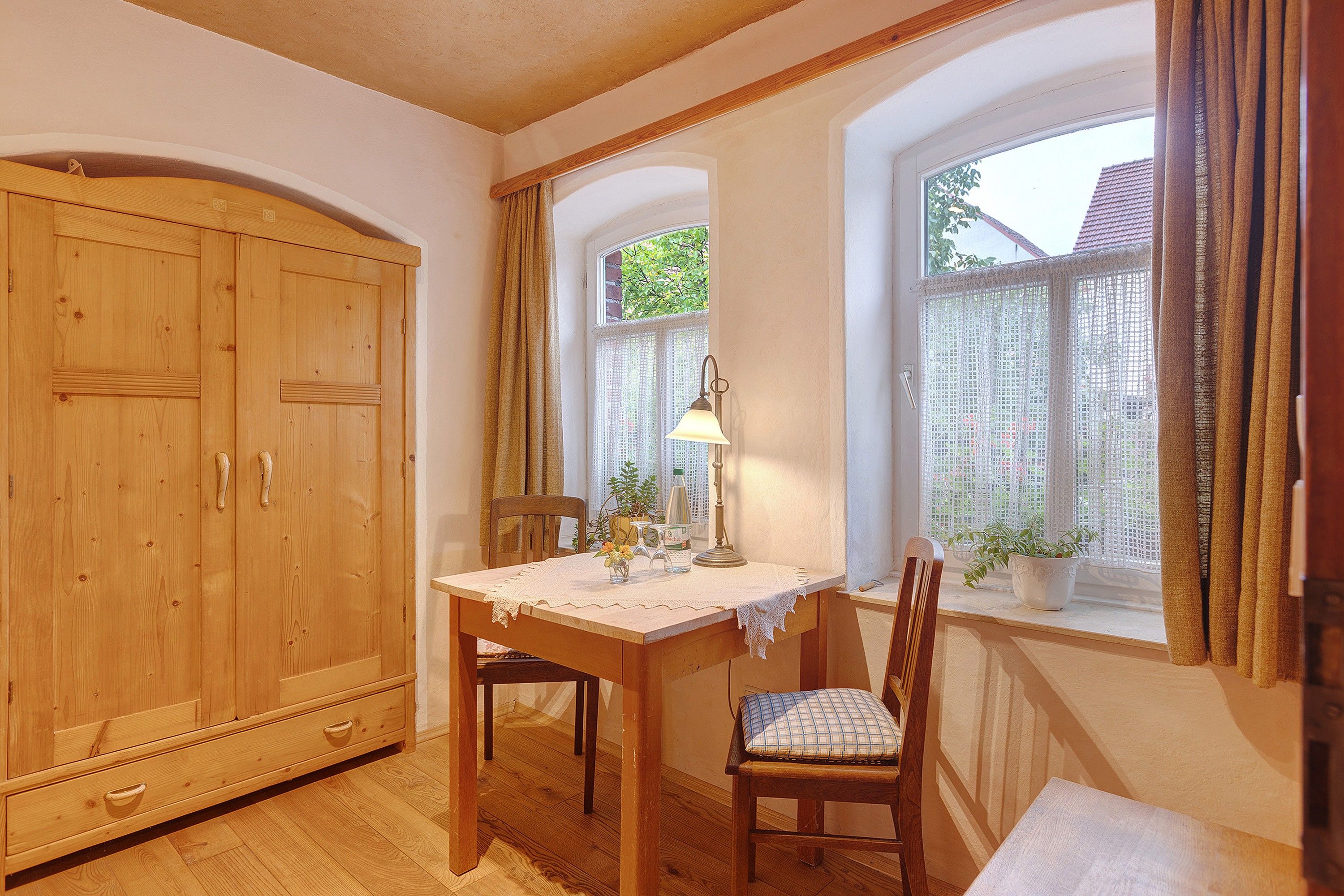 Ein Zimmer mit einem Holzschrank, einem Tisch mit Spitze, Stühlen, einer Lampe und einem Fenster mit Vorhängen.