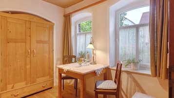 Ein Zimmer mit einem Holzschrank, einem Tisch mit Spitze, Stühlen, einer Lampe und einem Fenster mit Vorhängen.