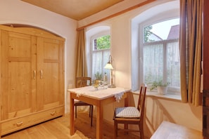 Une pièce comprenant une armoire en bois, une table recouverte d’une nappe en dentelle, des chaises, une lampe et une fenêtre avec des rideaux.
