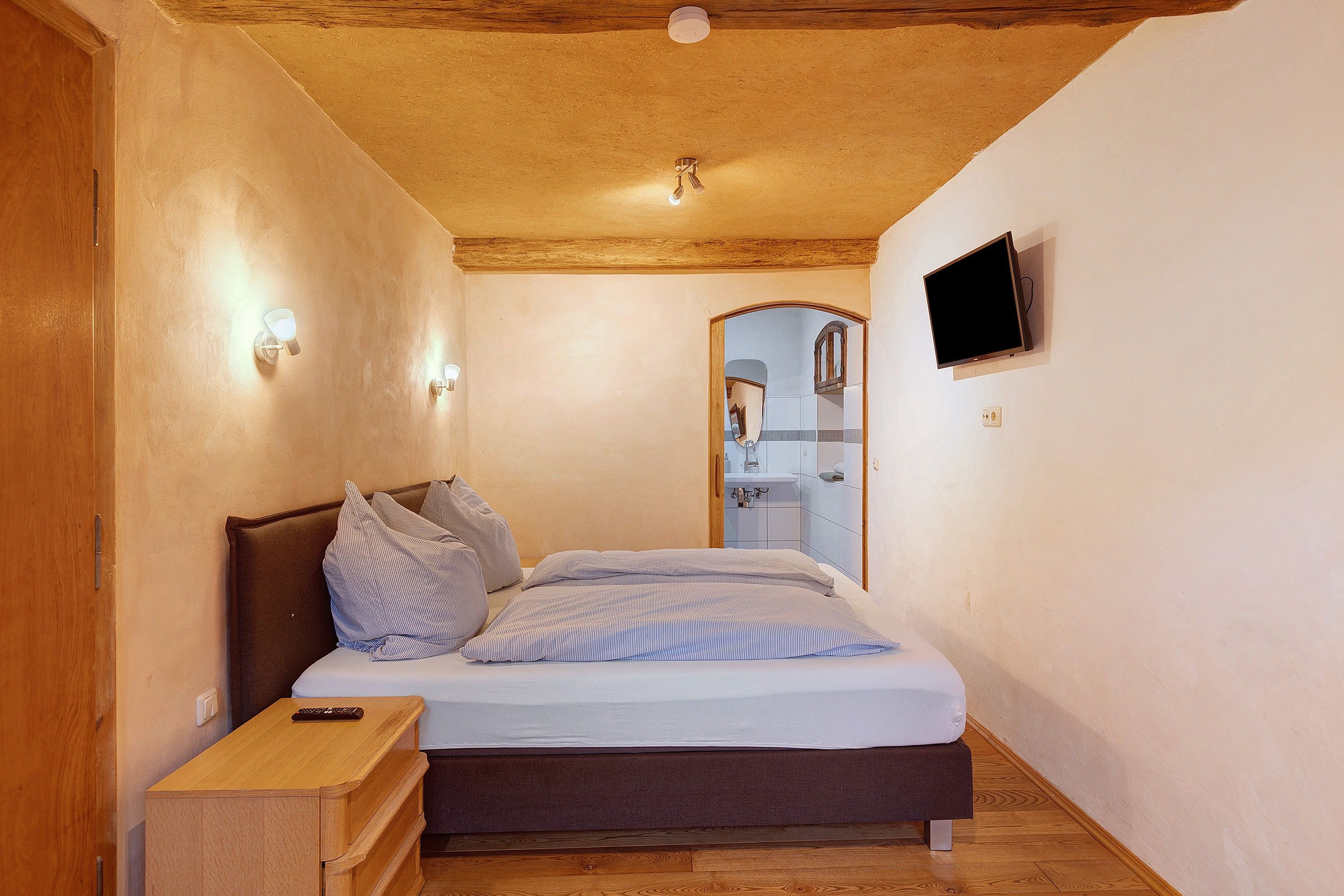 Ein Schlafzimmer mit Holzboden, einem Bett mit Kissen, einem Nachttisch, einem an der Wand montierten Fernseher und einem durch einen Bogen sichtbaren Badezimmer.