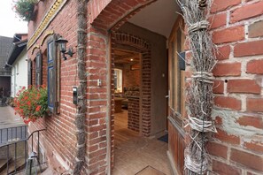 Un bâtiment en briques avec un escalier en bois menant à une porte, une plante suspendue et un espace intérieur visible, comprenant un canapé et une table.