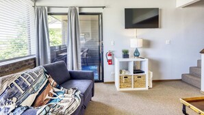 Ein Wohnzimmer mit einer Couch, einem Fernseher, einer Lampe, einem kleinen Tisch, einem Bücherregal und einem Feuerlöscher.