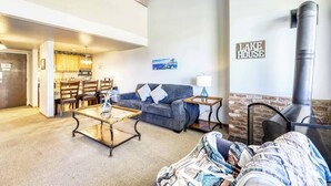 Ein Wohnzimmer mit Sofa, Couchtisch und Kamin. Es gibt eine Küchenzeile mit Holzausstattung und einem Esstisch. Ein Gemälde hängt an der Wand, und ein Schild zeigt 'LAKE HOUSE'.
