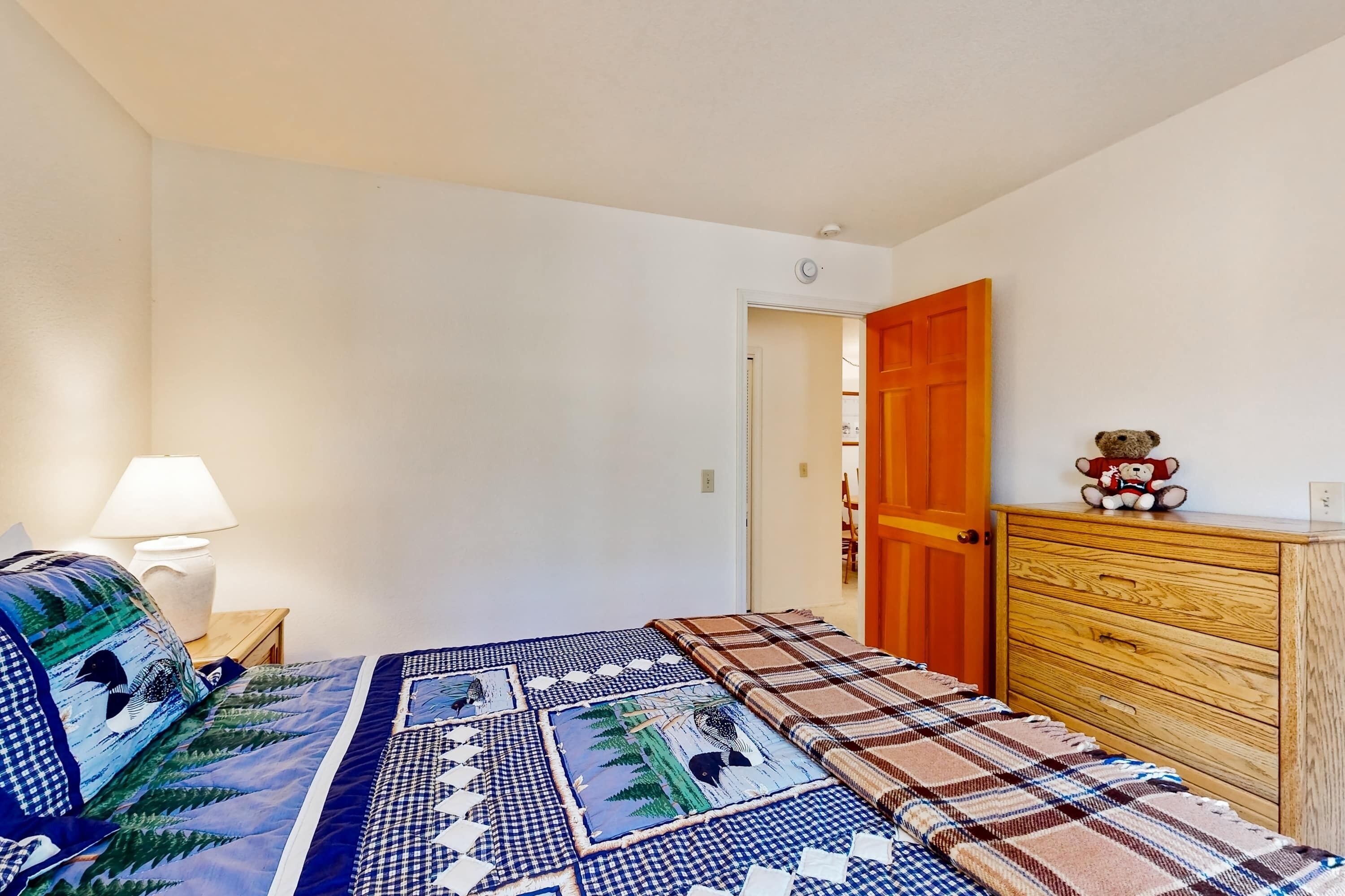 A bedroom with a bed, a nightstand, a wooden dresser, and a teddy bear on top.