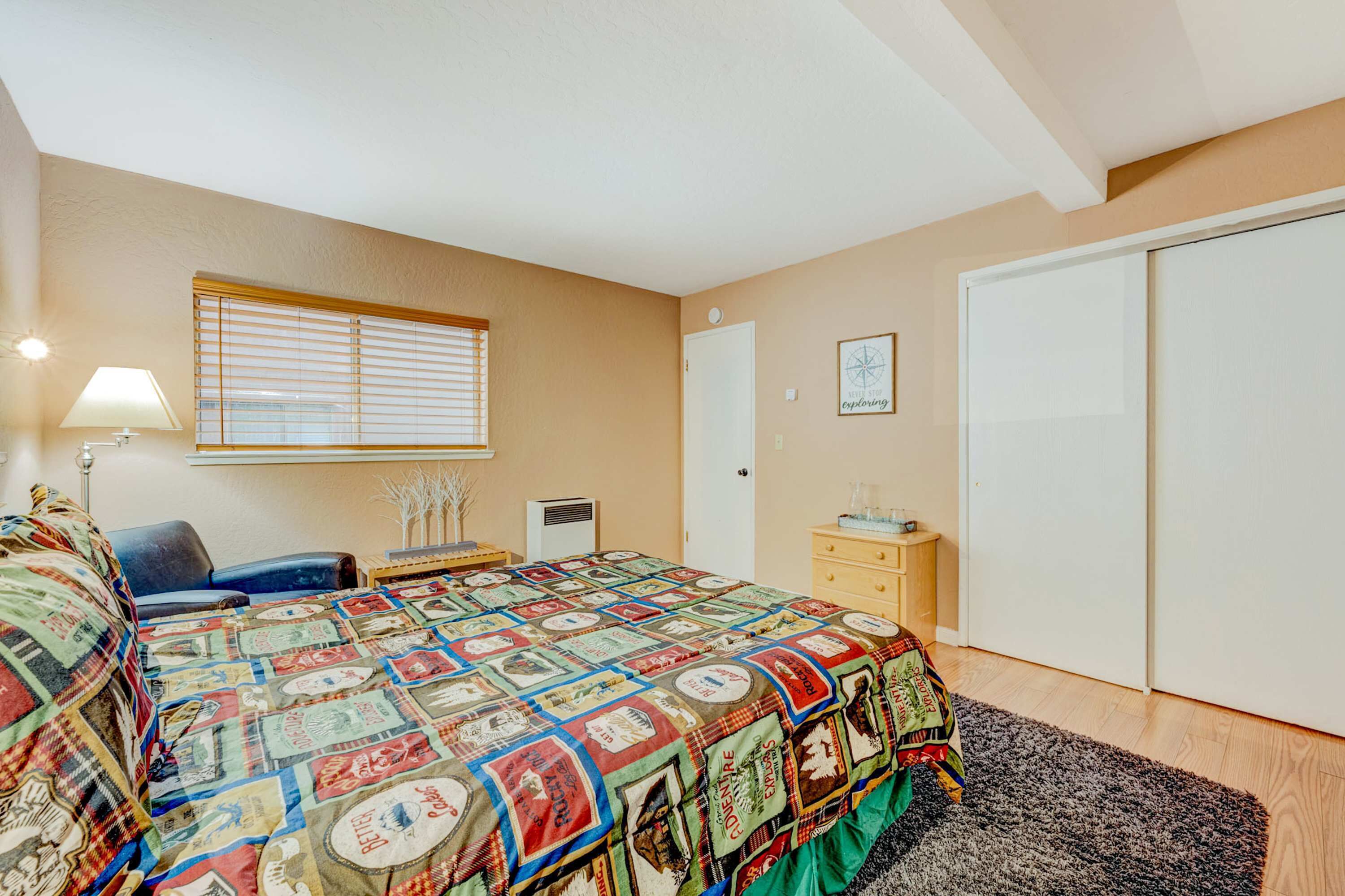A bedroom with a bed, a desk, a chair, a lamp, a window with blinds, a wardrobe, and a framed picture on the wall.
