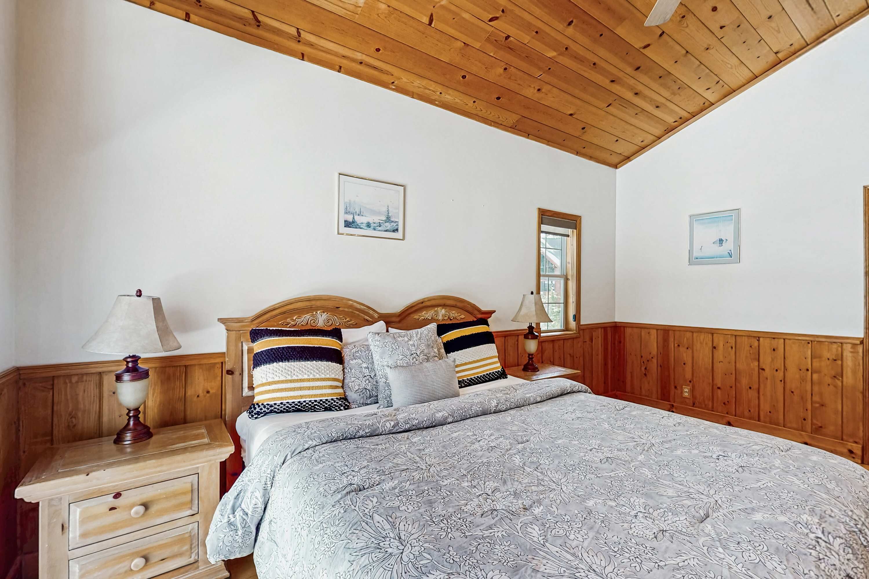 Ein Schlafzimmer mit Holzboden, einem Bett mit gemusterten Betttextilien, zwei Nachttischen mit Lampen und gerahmten Bildern an den Wänden.