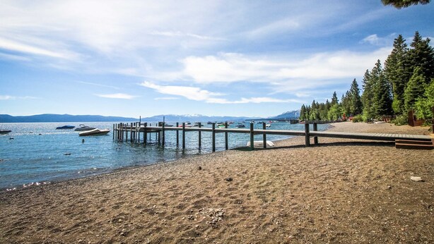Una playa con un muelle de madera que se adentra en aguas tranquilas, rodeada de árboles y montañas a lo lejos.