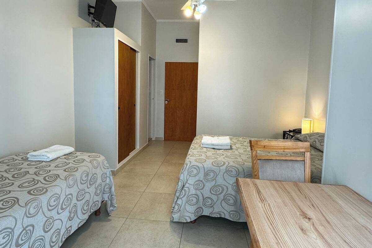 A hotel room with two single beds, a wooden table, a ceiling fan, and a TV mounted on the wall.