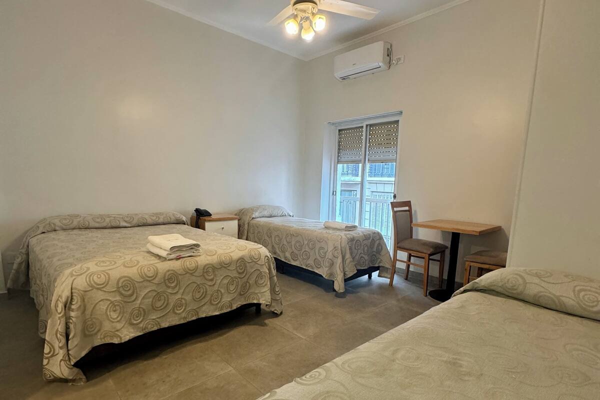 A hotel room with two beds, a ceiling fan, an air conditioning unit, a small table, and a window with a view.