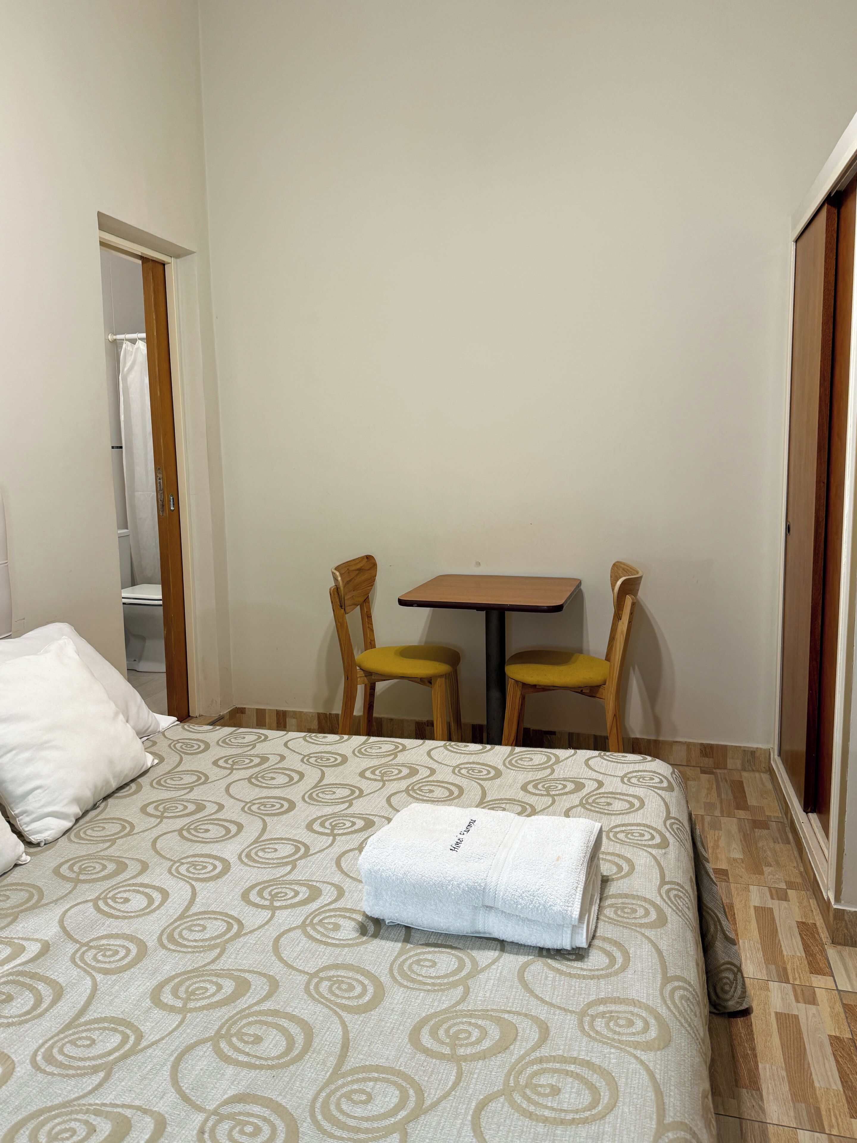 Quarto de hotel com cama, mesa e duas cadeiras, e porta de acesso a um banheiro.