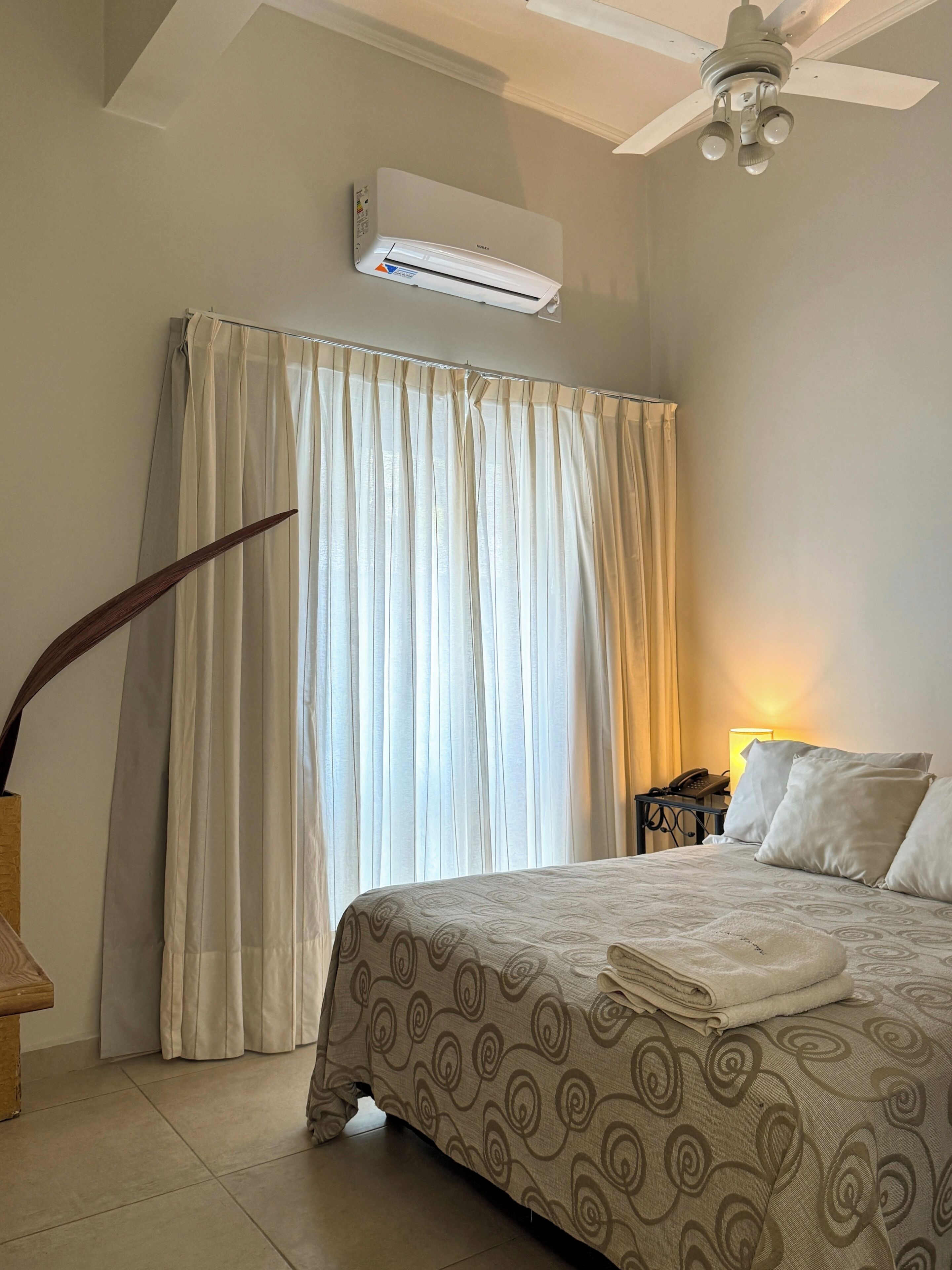 Et hotelværelse med seng, ventilator, aircondition, et natbord med lampe og telefon.