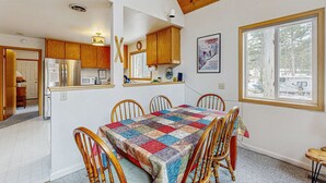 A dining area with a quilt-covered table, wooden chairs, a kitchen with wooden cabinets, a refrigerator, a microwave, and a window with a view of trees. - Cozy, dog-friendly getaway with wood stove & washer&dryer (Soda Springs)