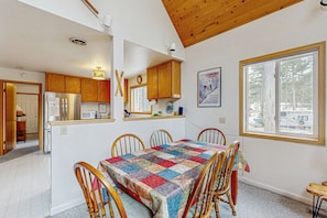 A dining area with a quilt-covered table, wooden chairs, a kitchen with wooden cabinets, a refrigerator, a microwave, and a window with a view of trees. - Cozy, dog-friendly getaway with wood stove & washer&dryer (Soda Springs)
