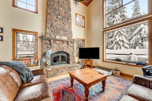 A cozy living room with a stone fireplace, leather sofas, a coffee table, and a large window with a snowy view.