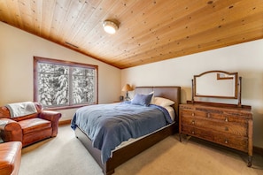 A bedroom with a wooden ceiling, a bed with a blue bedspread, a leather armchair, a dresser, and a window with a view of snow-covered trees.