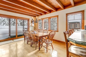 A dining area with a wooden table and chairs, large windows with views of snow-covered trees, and a chandelier.