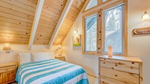 A bedroom with a sloped ceiling, a bed with striped bedding, a wooden dresser, a nightstand, a wall-mounted lamp, a window with a view of trees, and a wall decoration. - close to beach & skiing with grill, fireplace, deck, & W&D (Tahoma)