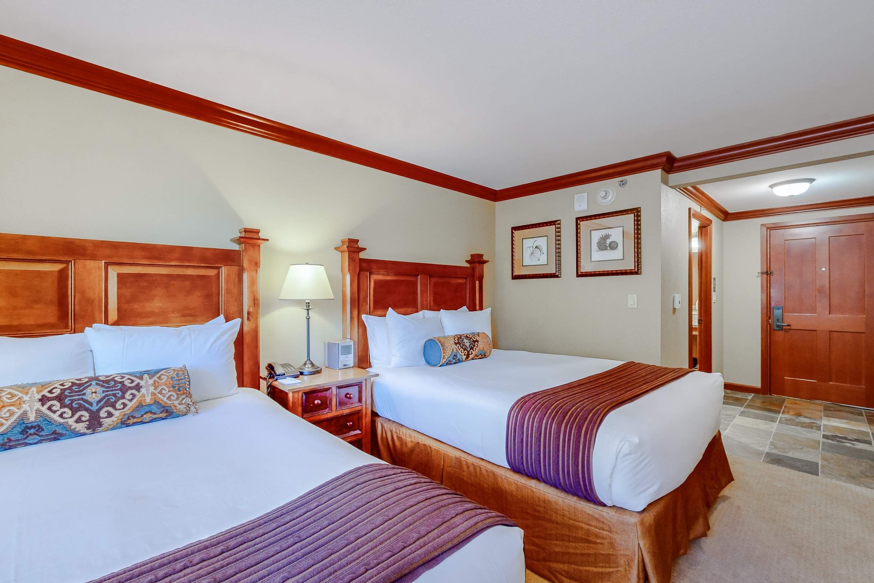 A hotel room with two beds, wooden headboards, a nightstand with a lamp, and a door leading to another room.