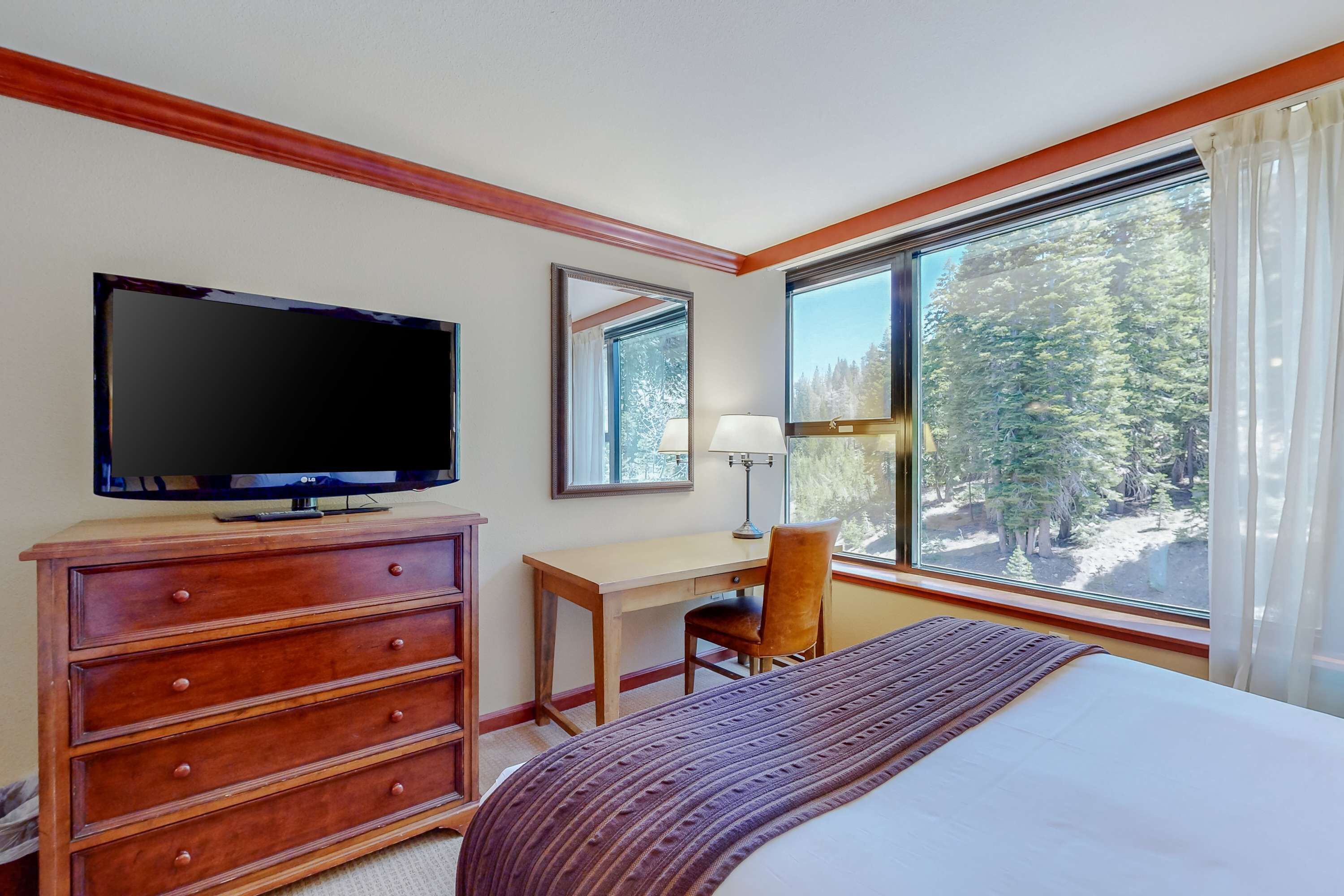 Une chambre d’hôtel équipée d’un lit, d’un bureau, d’une télévision, d’une chaise et d’une fenêtre donnant sur la verdure.