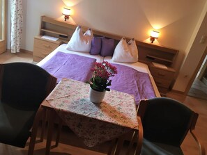 Ein Hotelzimmer mit einem Bett, zwei Nachttischlampen, einem Nachttisch, einem Tisch mit einer blumigen Tischdecke, zwei Stühlen und einer Topfpflanze.