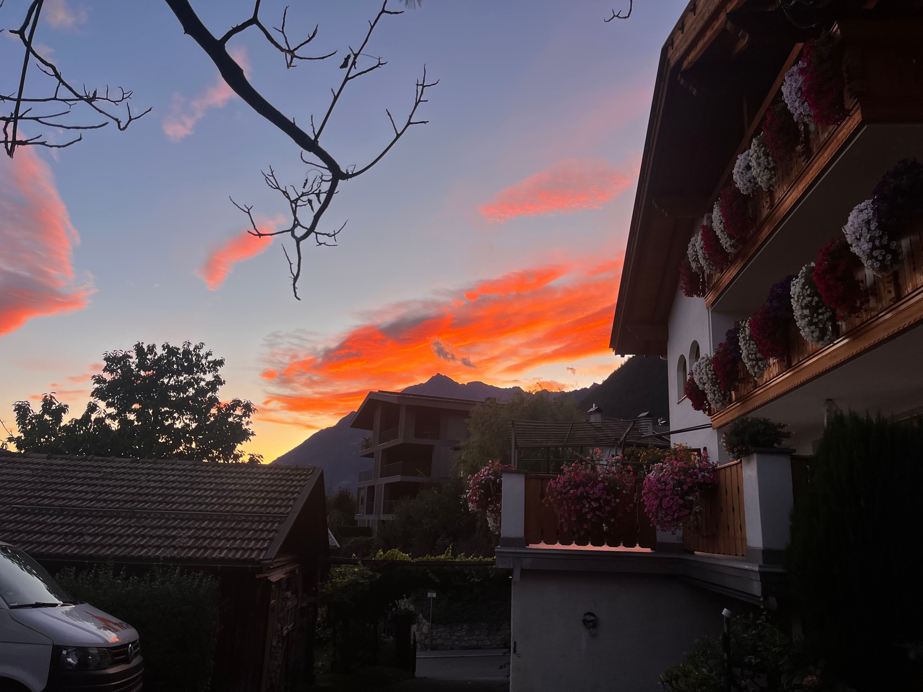 Ein Wohngebiet mit einem Haus, das einen Balkon mit Blumenkästen, ein geparktes Auto davor und einen Berg im Hintergrund zeigt.