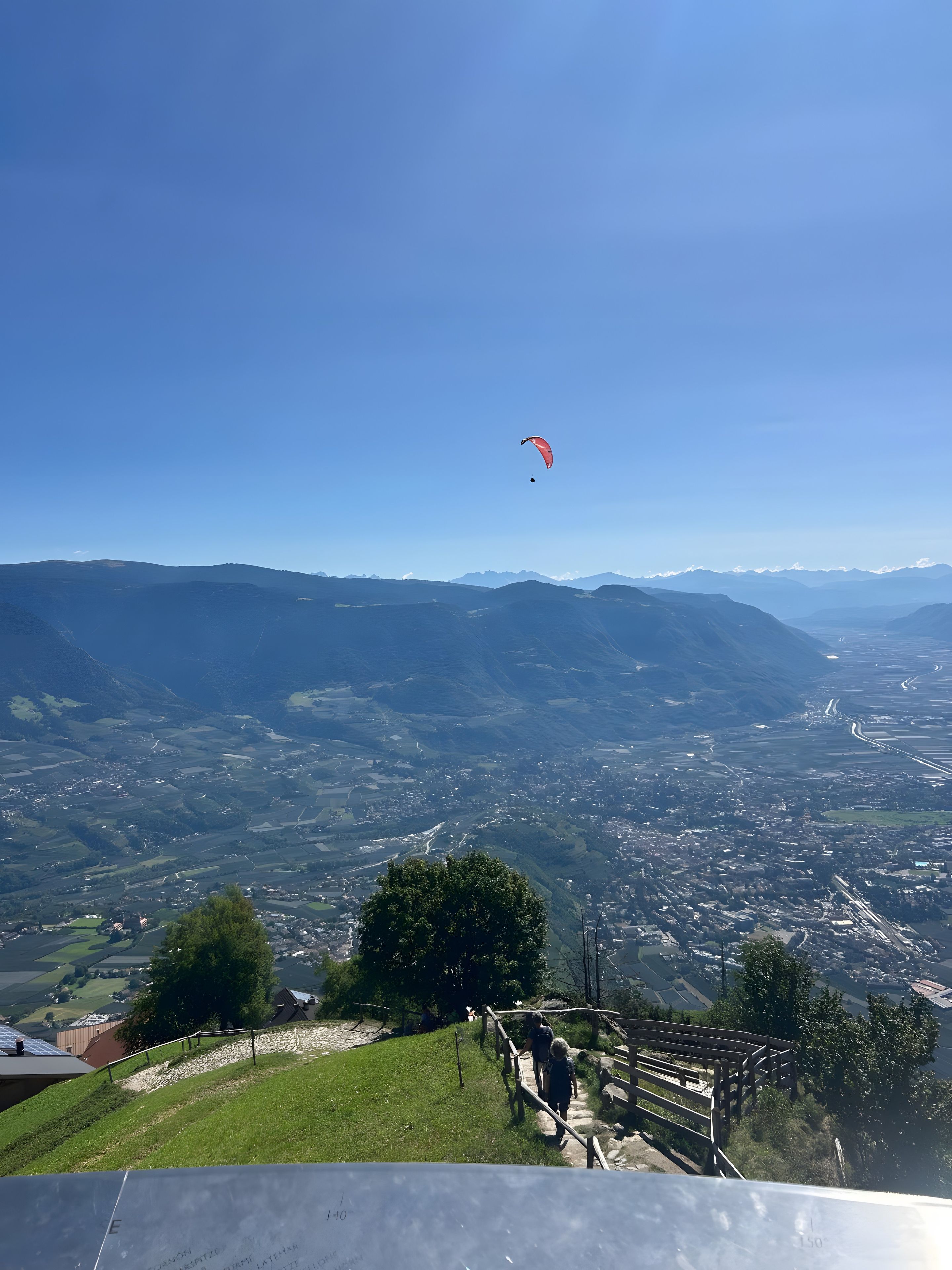 Vista panoràmica d'un paisatge muntanyós, amb un parapent vermell al cel, un camí sinuós i un cel blau clar.