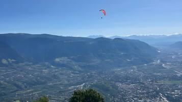 Vista panoràmica d'un paisatge muntanyós, amb un parapent vermell al cel, un camí sinuós i un cel blau clar.
