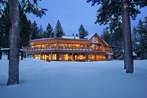 Un grand chalet à plusieurs étages, doté d’un porche couvert et de nombreuses fenêtres, entouré de neige et d’arbres.