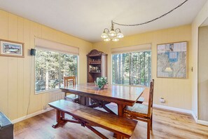 Ein Essbereich mit Holztisch, Stühlen, einer Chandelüster und großen Fenstern mit Blick auf Bäume.