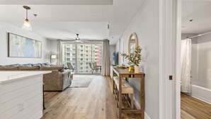 A modern living room with a sofa, a dining table, and a large window with a view of high-rise buildings. - Shores of Panama 1416~2 Bdrm~sleeps 6 (Panama City Beach)