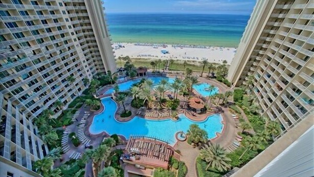 A high-rise hotel with a central swimming pool surrounded by palm trees and a beach view. - Shores of Panama 1416~2 Bdrm~sleeps 6 (Panama City Beach)