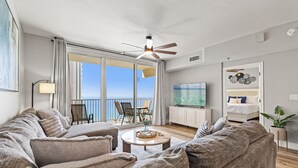 A living room with a large sofa, a coffee table, and a TV. There is a balcony with a view of the sea. - Shores of Panama 1416~2 Bdrm~sleeps 6 (Panama City Beach)