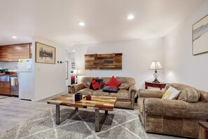 A living room with a sofa, coffee table, and a kitchen in the background. - All-season cabin with wraparound deck - quick walk to the beach (Carnelian Bay)