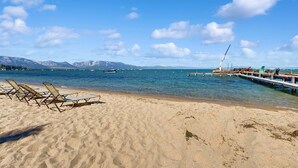A beach with sun loungers, a pier extending into the water, and mountains in the background.
