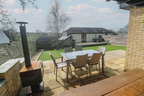 A patio with a table and chairs, a barbecue grill, and a view of a house and garden. - Ash Barn (Falmouth)