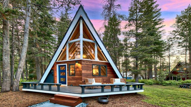 A cabin with a triangular roof, a deck with seating, and a view of the surrounding forest. - Beautiful A-frame home with deck, streaming, and dog friendly - near skiing (South Lake Tahoe)