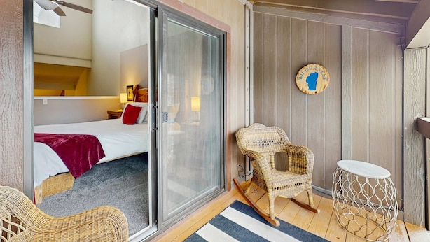 A room with a bed, a wicker chair, a small round table, and a sliding glass door leading to another room.